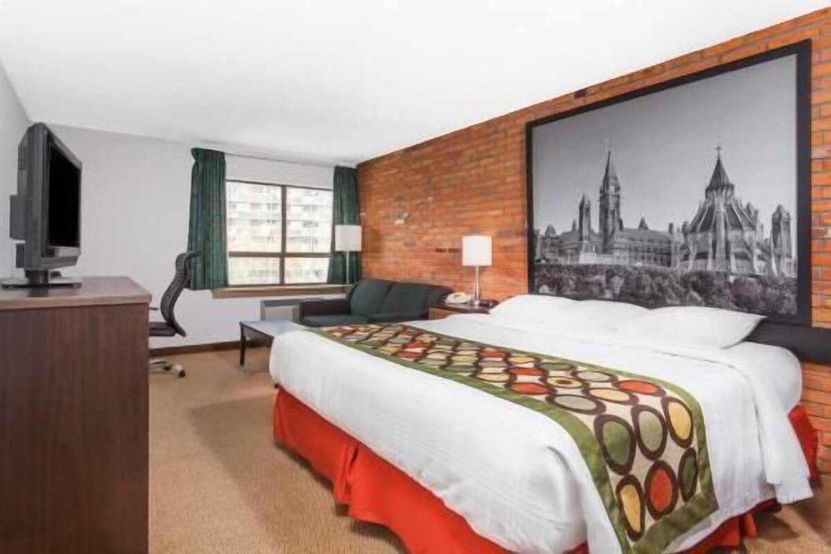 Une chambre d’hôtel avec un lit, une télévision, un bureau, une chaise, une fenêtre et un tableau représentant un paysage urbain accroché au mur.