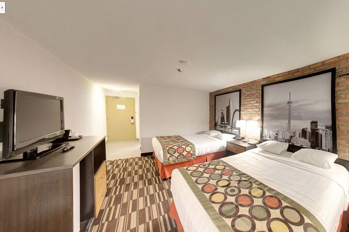 Une chambre d’hôtel avec deux lits, une télévision, une fresque représentant un paysage urbain et un mur en briques.