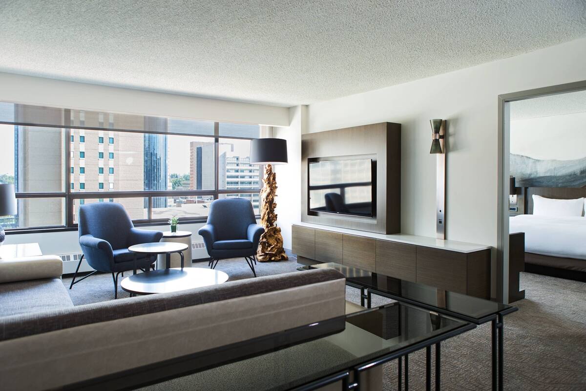 Una habitación de hotel moderna con sofá, dos sillones, una mesa de centro, televisión, cama y un ventanal con vistas a la ciudad.