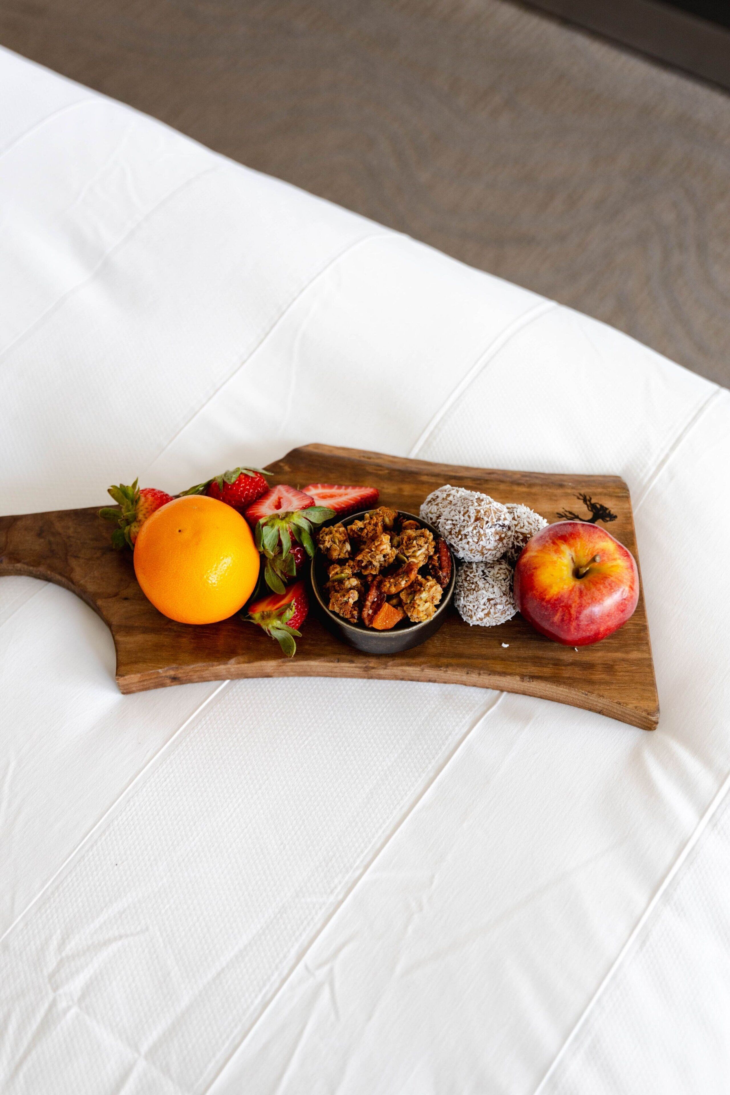 A wooden serving board with fruits and a bowl of granola on a white fabric surface.