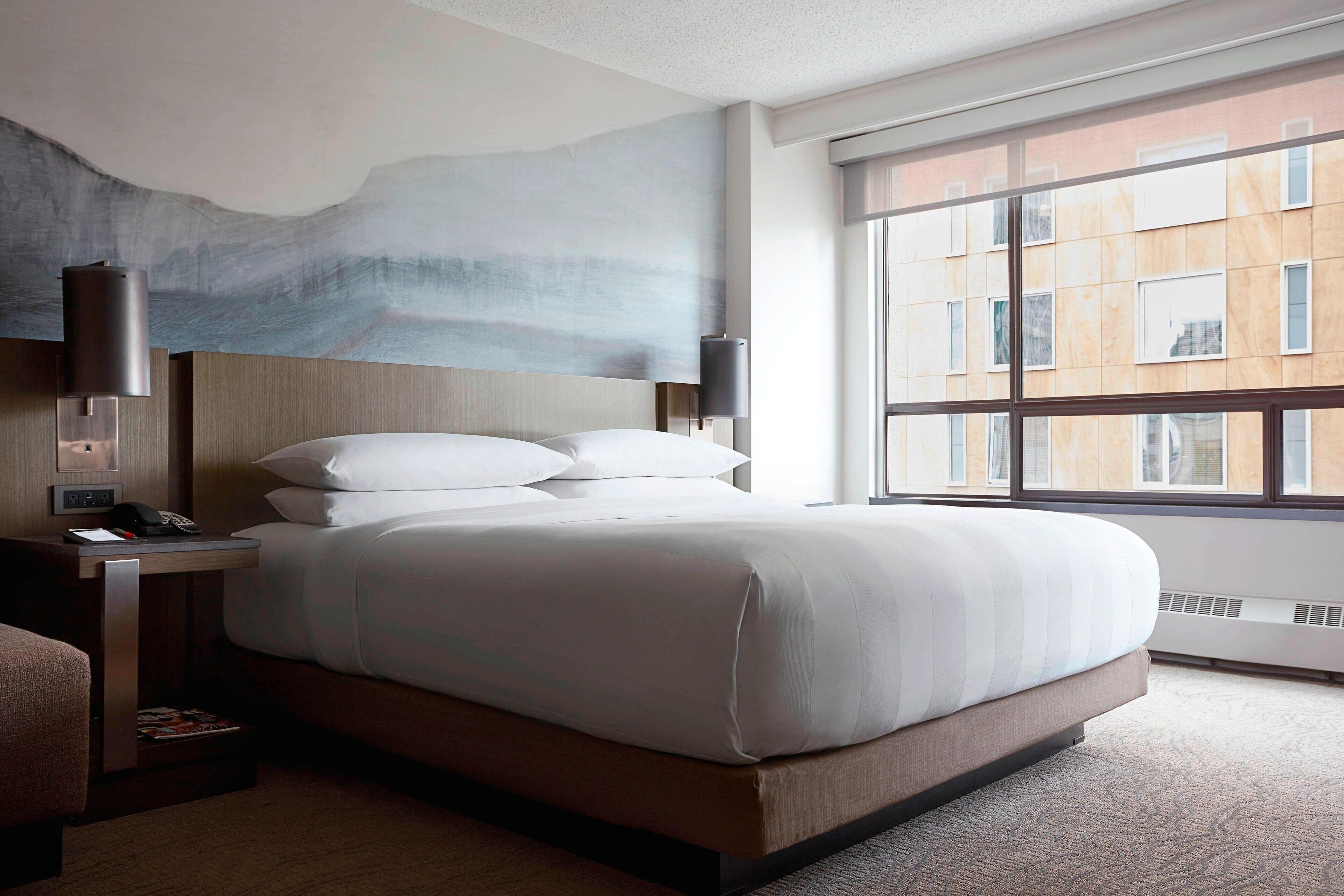 Una habitación de hotel moderna con una cama grande, una mesita de noche, una lámpara y vistas de la ciudad a través de amplios ventanales.