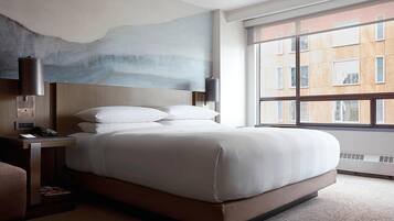 Una habitación de hotel moderna con una cama grande, una mesita de noche, una lámpara y vistas de la ciudad a través de amplios ventanales.