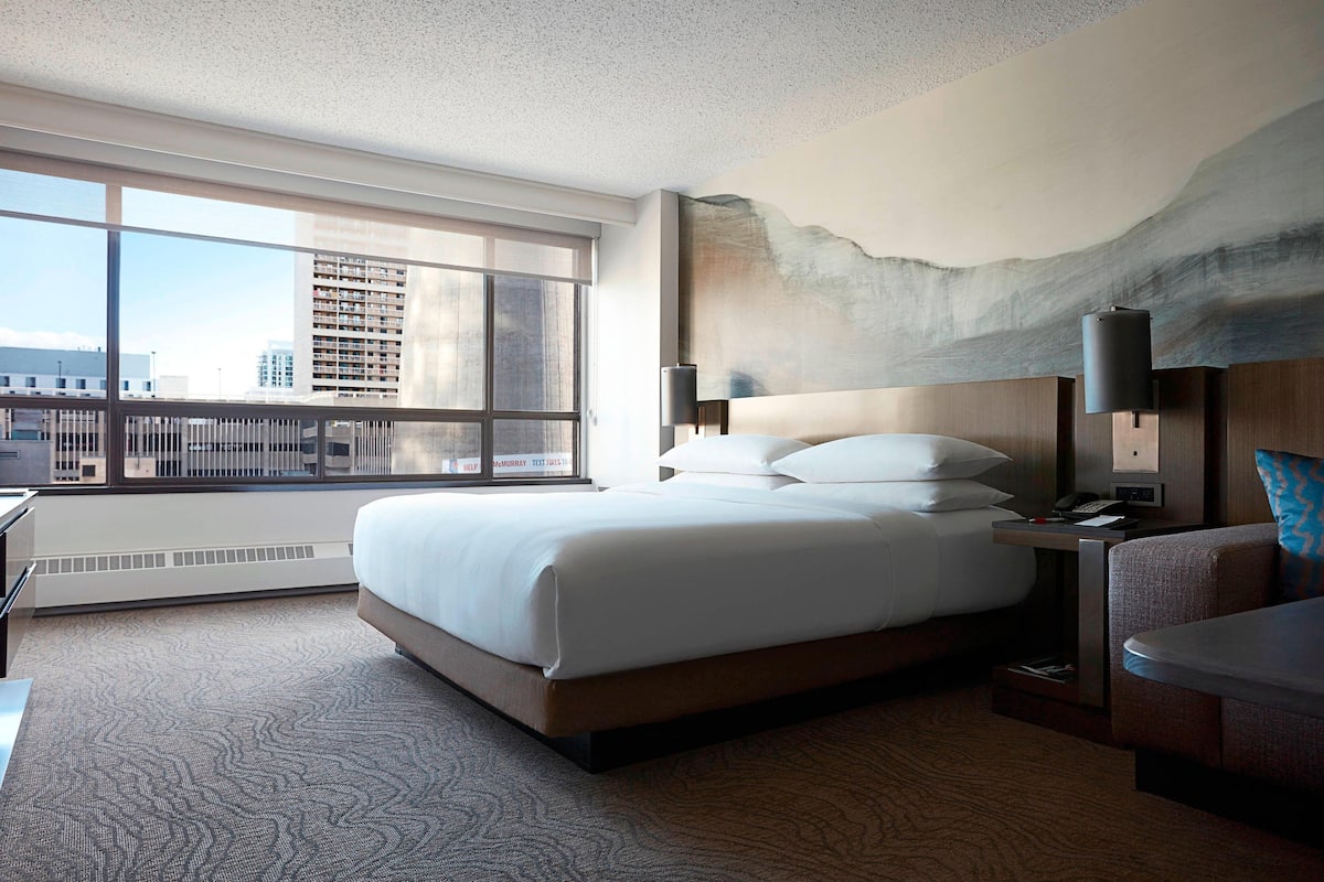 Habitación de hotel con una cama grande, un escritorio, una silla y vistas a la ciudad a través de amplias ventanas.