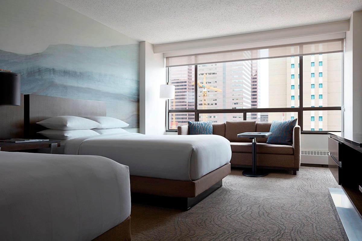 Habitación de hotel con una cama grande, un sofá, una mesita y un televisor. Se ve una ventana con vistas a edificios.