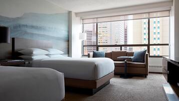 Habitación de hotel con una cama grande, un sofá, una mesita y un televisor. Se ve una ventana con vistas a edificios.