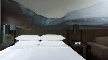 Une chambre d’hôtel avec un grand tableau abstrait au mur, un lit avec du linge de maison blanc, une table de chevet avec une lampe et une chaise.
