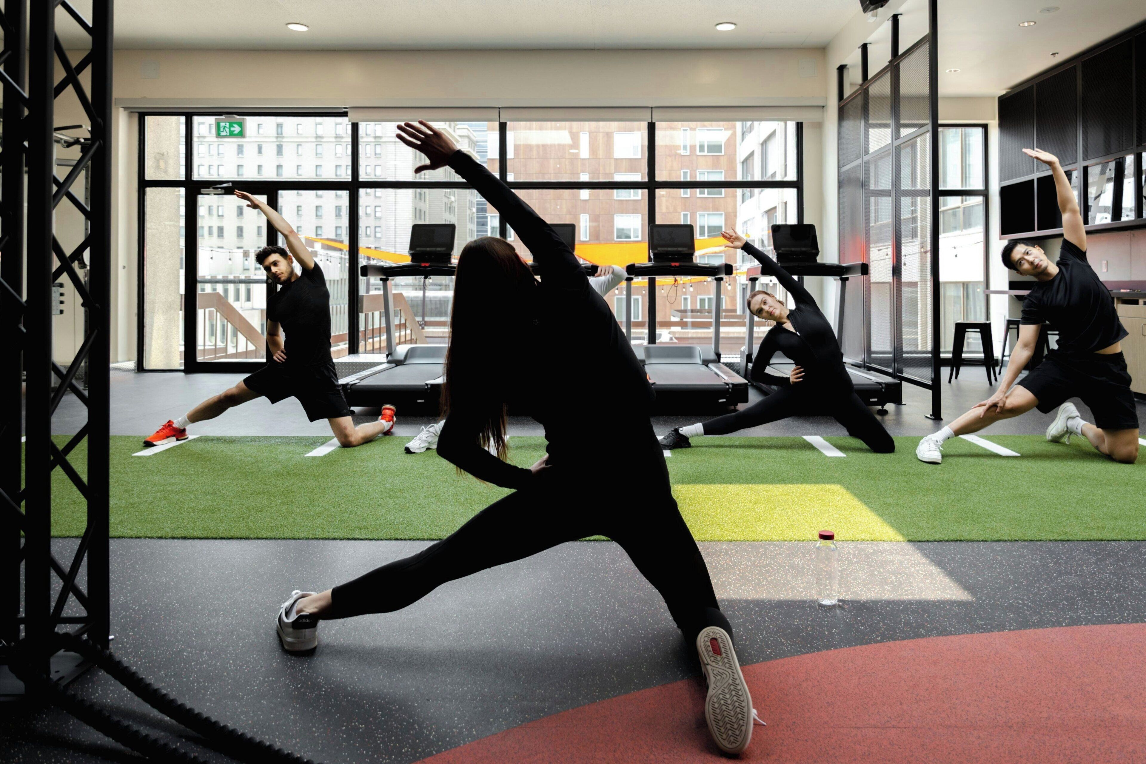 Un grupo de personas realizan ejercicios de estiramiento en un gimnasio con amplias ventanas y equipo de entrenamiento.