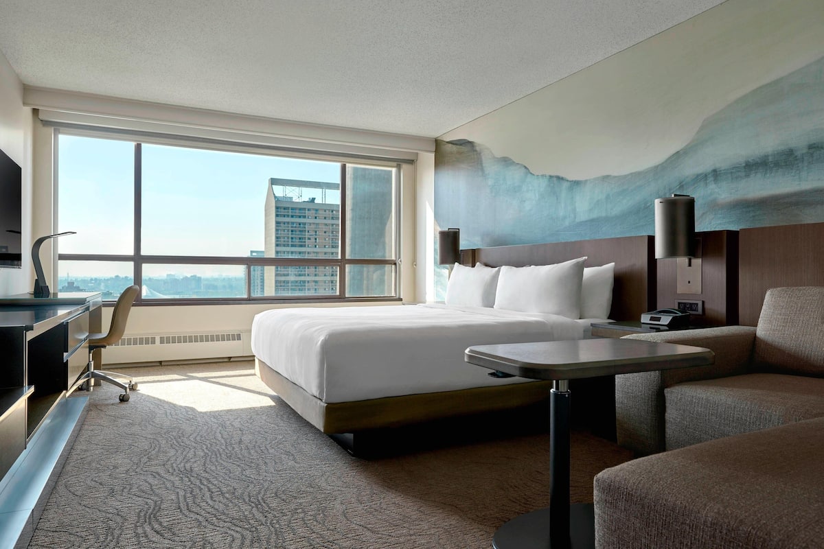 Habitación de hotel con una cama grande, un escritorio, una silla, un sofá y vistas de la ciudad a través de amplios ventanales.