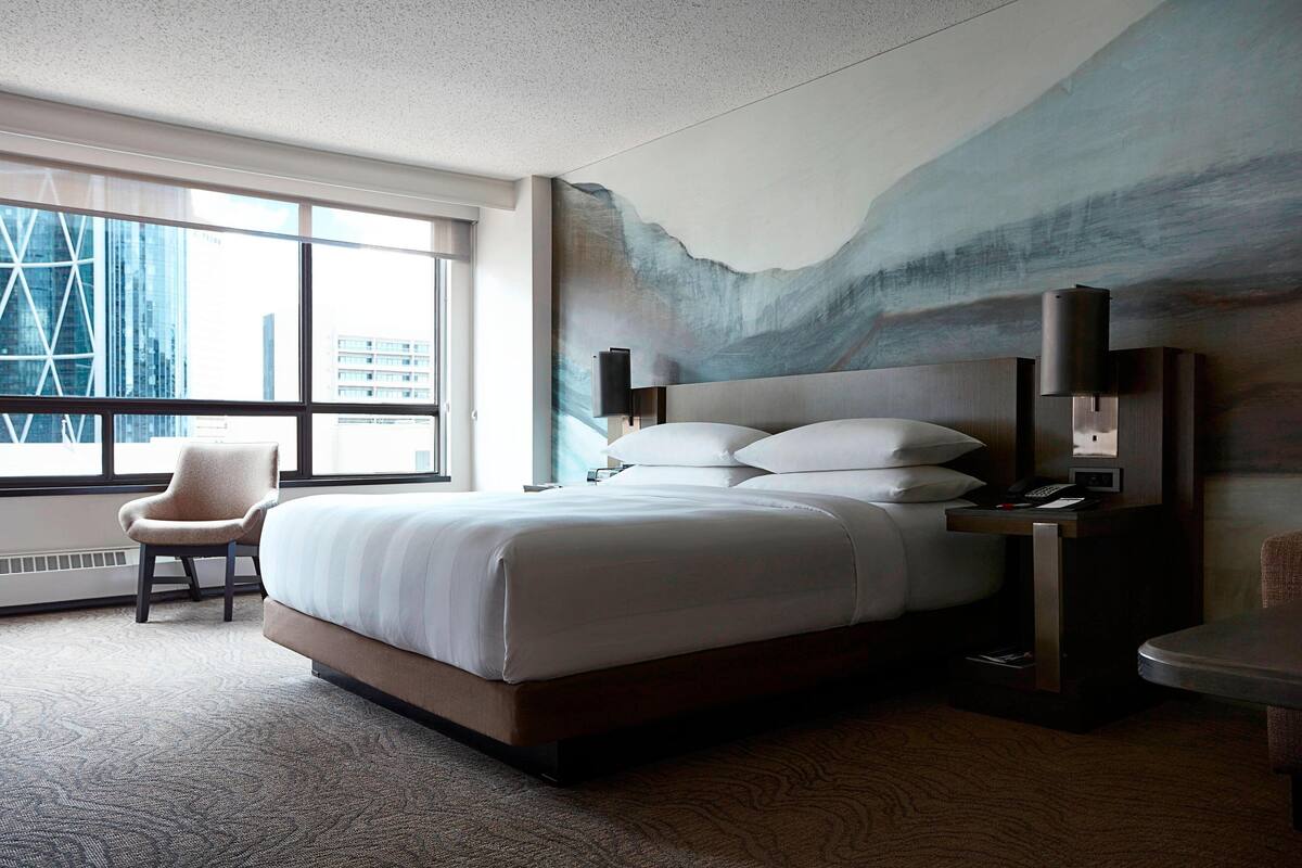 Habitación de hotel con una cama grande, una silla, un escritorio y vistas de la ciudad a través de amplios ventanales.