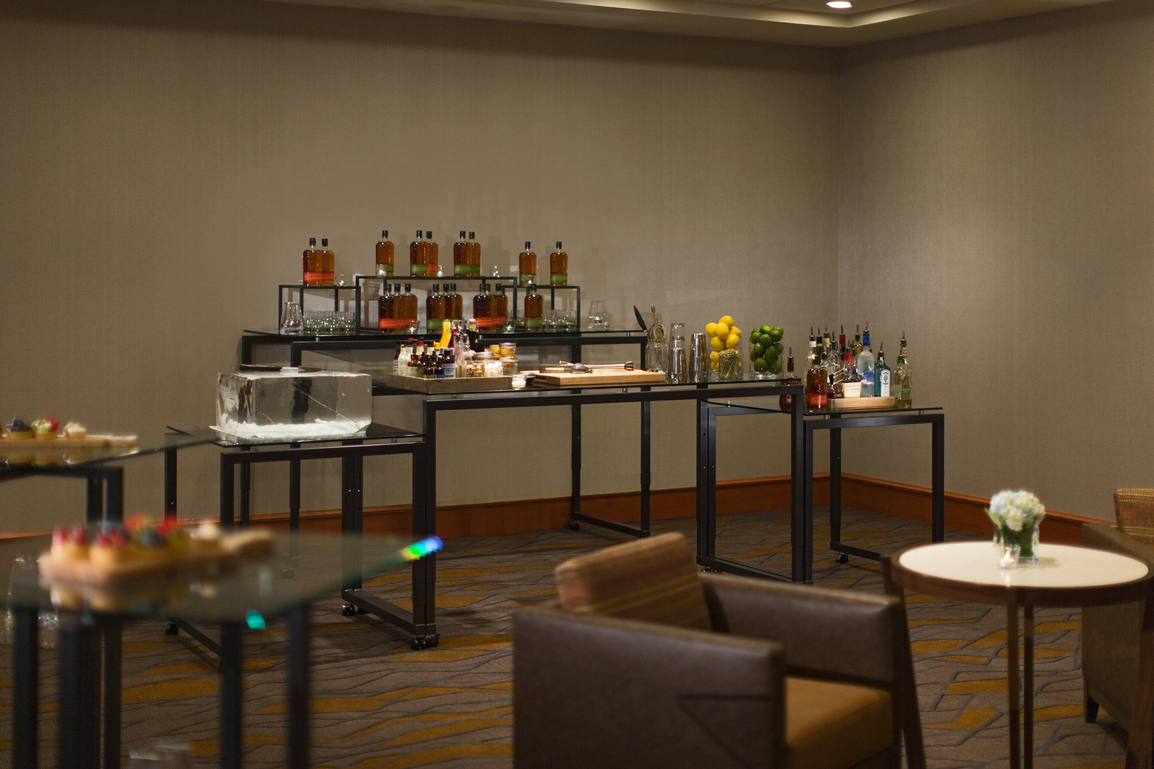 Un hall de hotel con zona de bar que muestra diversas botellas y vasos, una sala de estar con sillas y mesas, y suelo alfombrado.