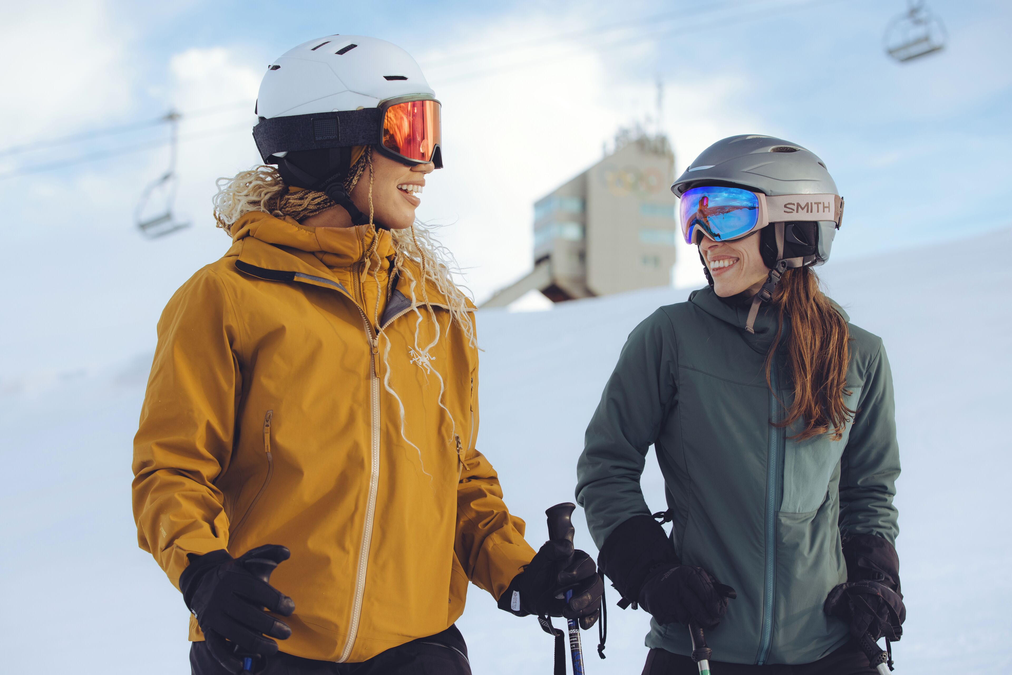 Dos personas con equipo de esquí de pie en una pendiente nevada, con telesillas al fondo.