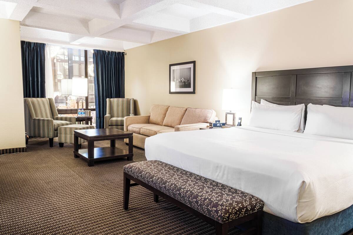 A hotel room with a large bed, a sofa, two armchairs, a small table, and a bench.