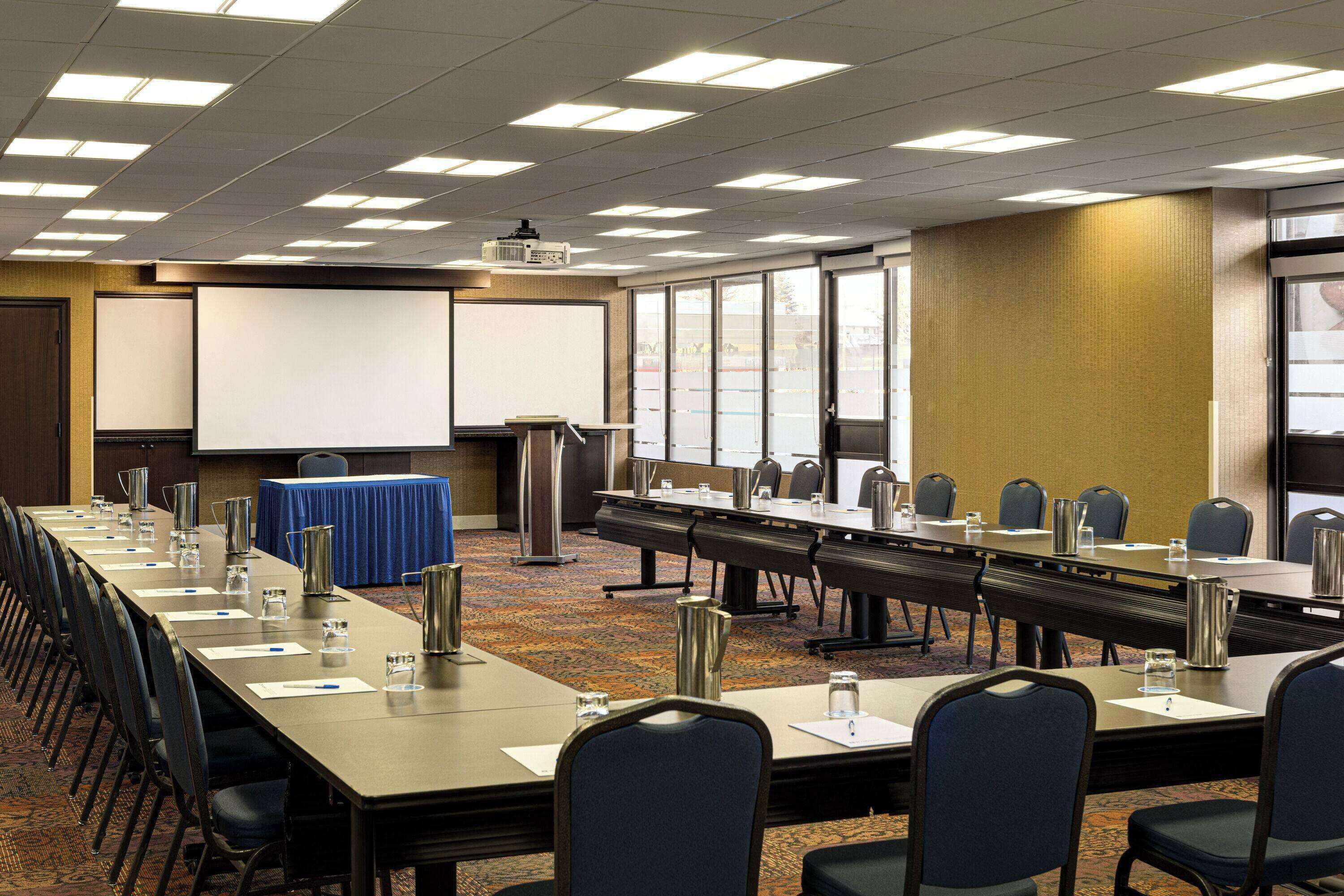 Un salón de conferencias con una larga mesa, sillas, una pantalla de proyector y un mantel azul.