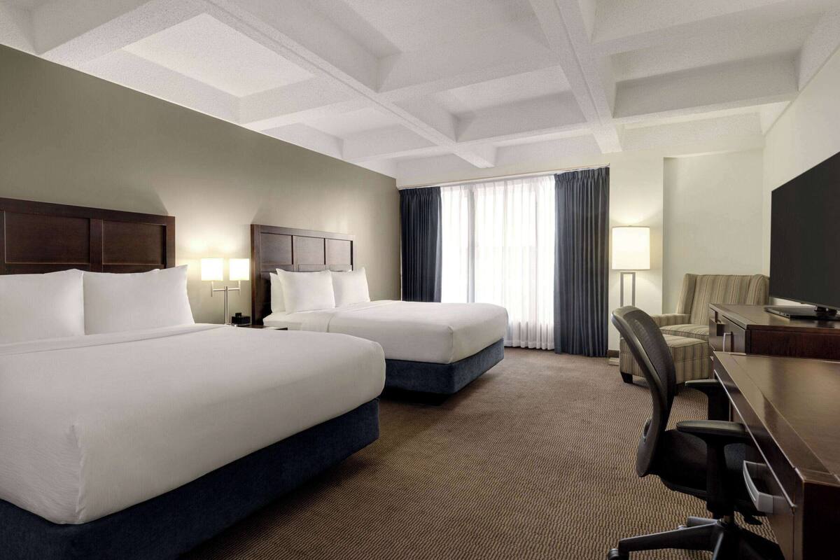 A hotel room with two beds, a desk, a chair, a TV, and a window with curtains.