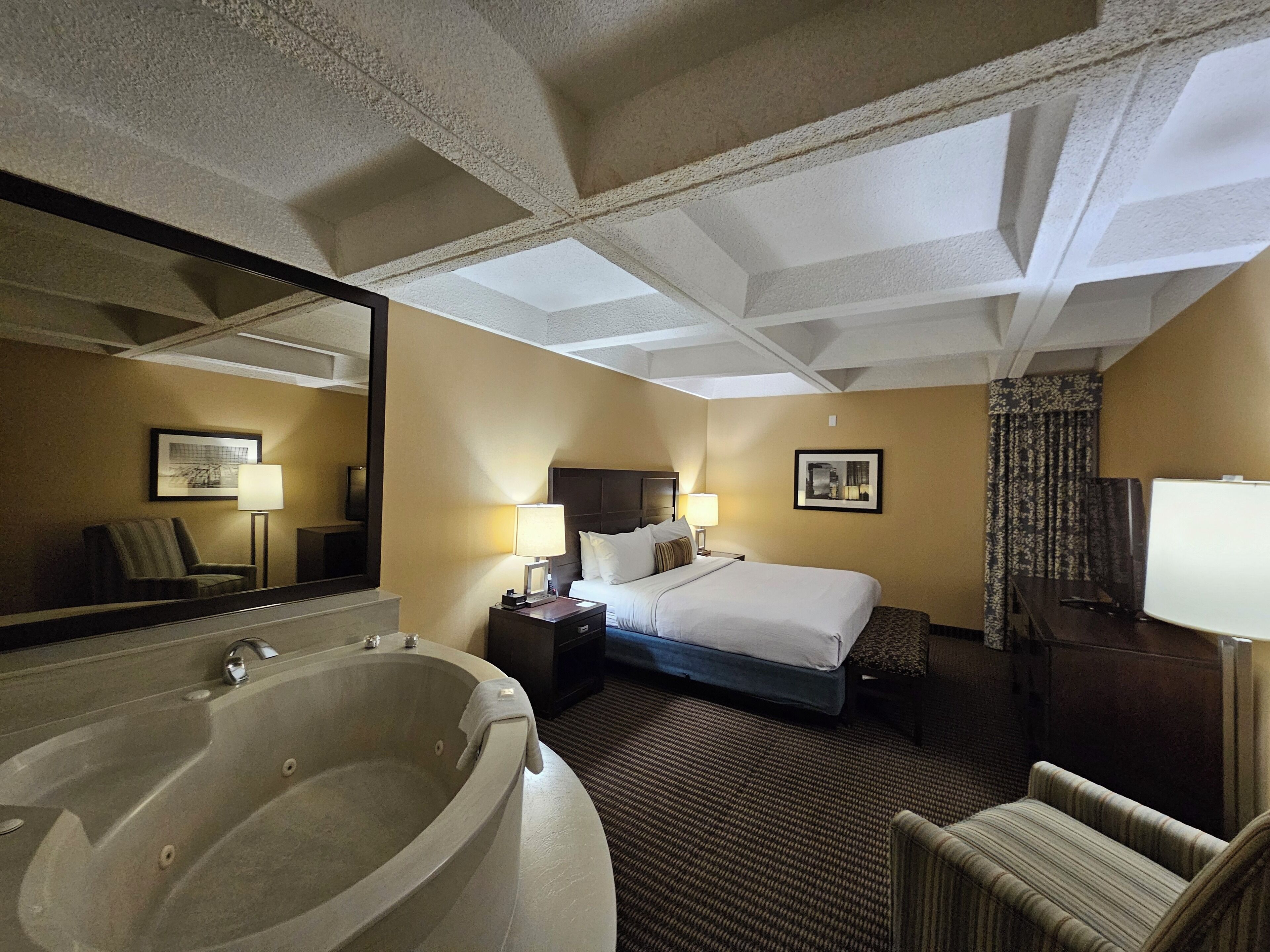 Ein Hotelzimmer mit einer großen Badewanne, einem Bett mit weißen Laken, einem Nachttisch mit Lampen und einem Spiegel an der Wand.