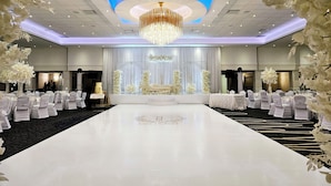 Un salón para recepción de bodas con alfombra blanca, un escenario central con decoración floral y una araña.