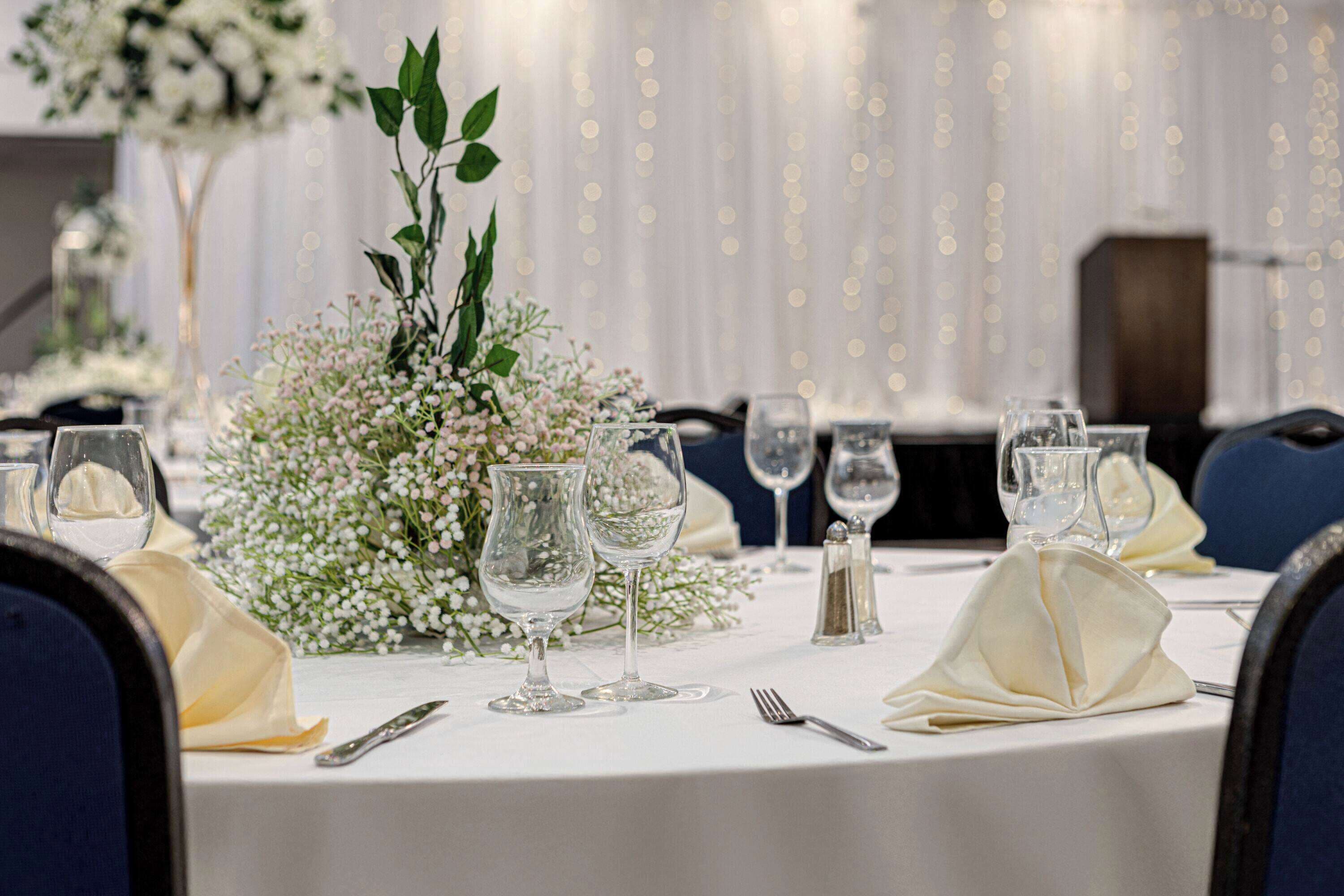 A table set for a formal event with white tablecloths, clear glassware, and a centerpiece of white flowers and greenery.