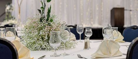 A table set for a formal event with white tablecloths, clear glassware, and a centerpiece of white flowers and greenery.