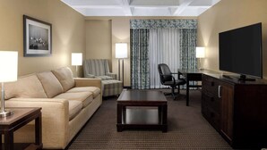 Habitación de hotel con sofá, sillón, escritorio, televisor y un amplio ventanal con cortinas.