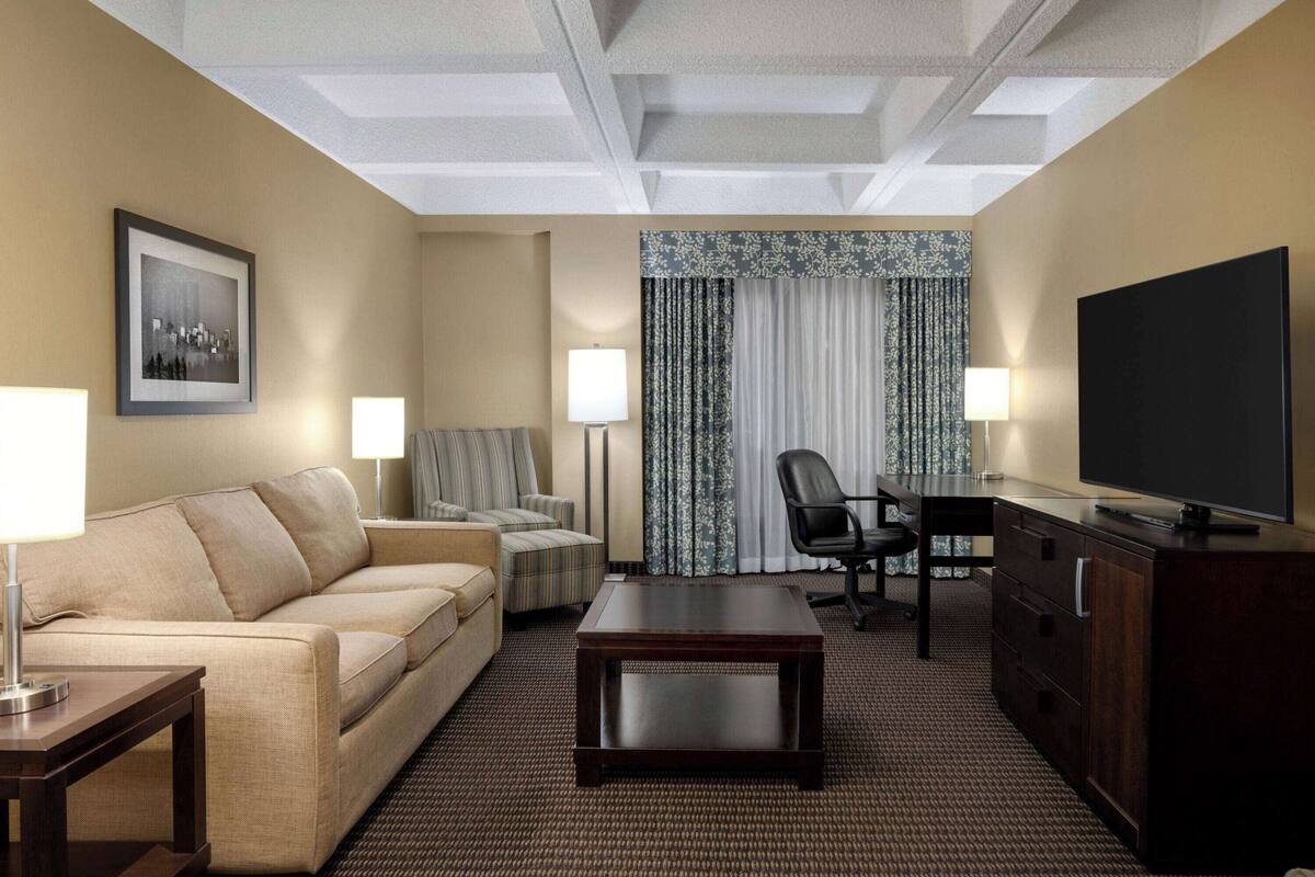 A hotel room with a sofa, armchair, desk, television, and a large window with curtains.