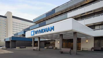 Uma entrada de hotel moderna com um letreiro de 'WYNDHAM' em destaque, área de estar coberta e um edifício de vários andares ao fundo.
