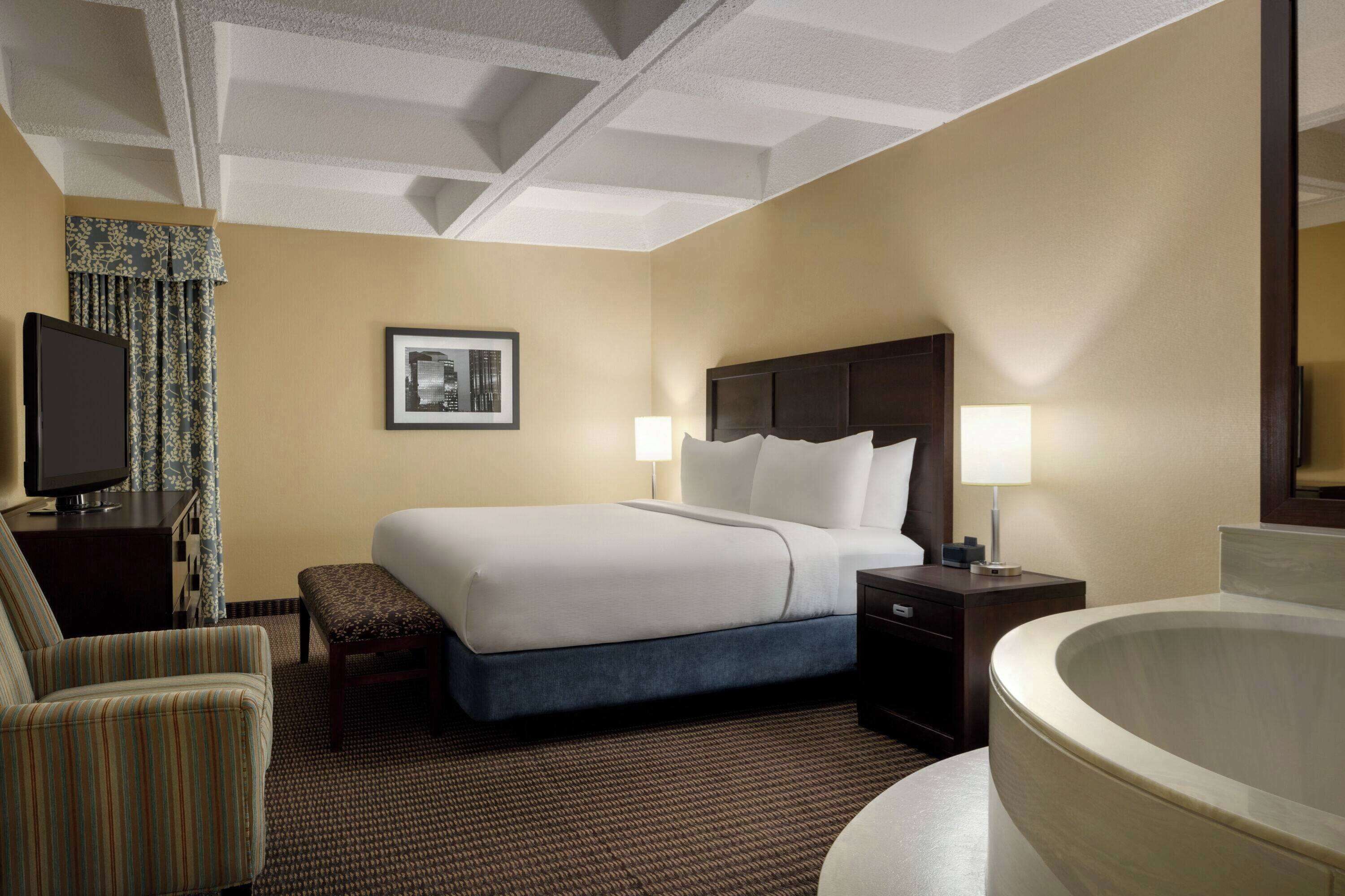 A hotel room with a large bed, a desk with a television, a chair, a bench, a bedside table with lamps, and a bathtub.