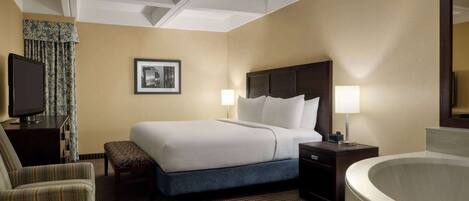 A hotel room with a large bed, a desk with a television, a chair, a bench, a bedside table with lamps, and a bathtub.