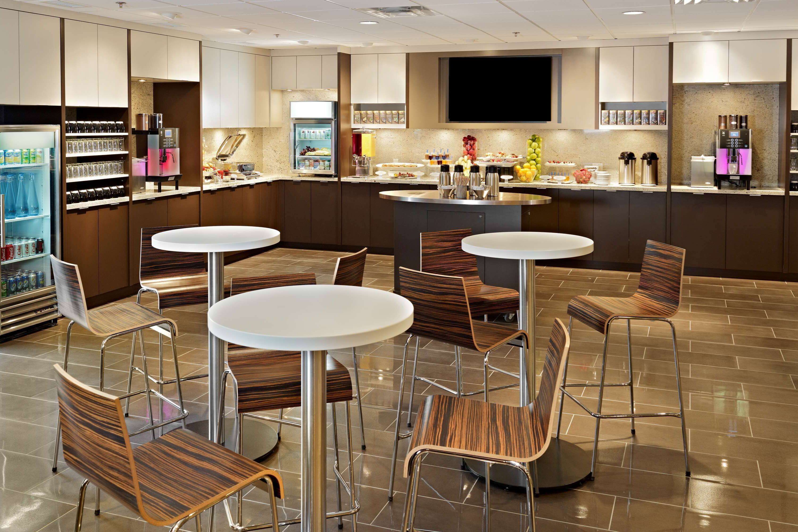 A modern hotel lobby with a central bar area, seating arrangements, and a self-service beverage station.