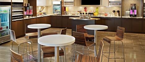 A modern hotel lobby with a central bar area, seating arrangements, and a self-service beverage station.