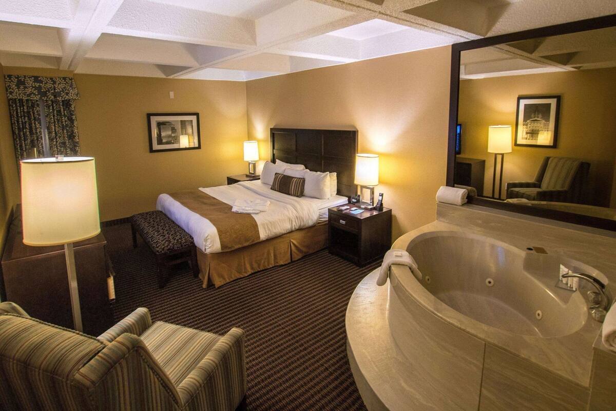 A hotel room with a bed, bedside lamps, a chair, a large mirror, and a bathtub.