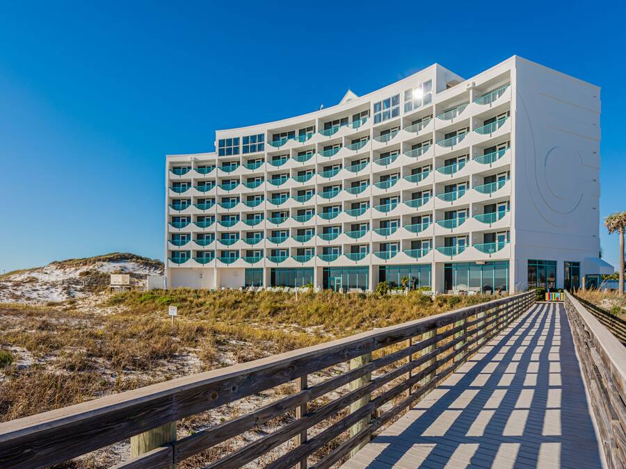 A modern hotel with a multi-story structure, a wooden boardwalk leading to the entrance, and a clear blue sky.