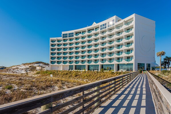A modern hotel with a multi-story structure, a wooden boardwalk leading to the entrance, and a clear blue sky.