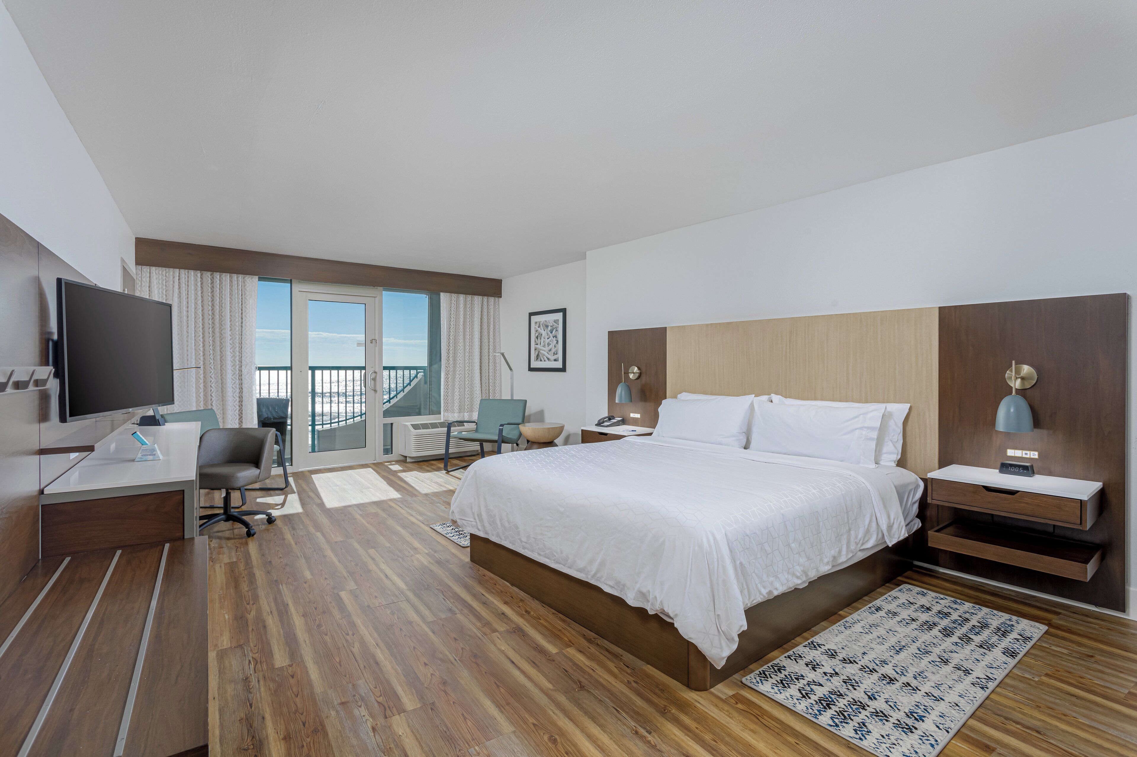 Habitación de hotel con una cama grande, un escritorio con televisor, una silla, una mesita de noche con lámpara y vistas al exterior.