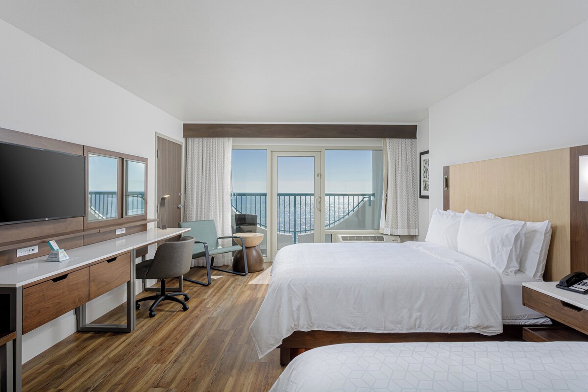 Habitación de hotel con cama, escritorio, silla, televisor y balcón con vista al mar.