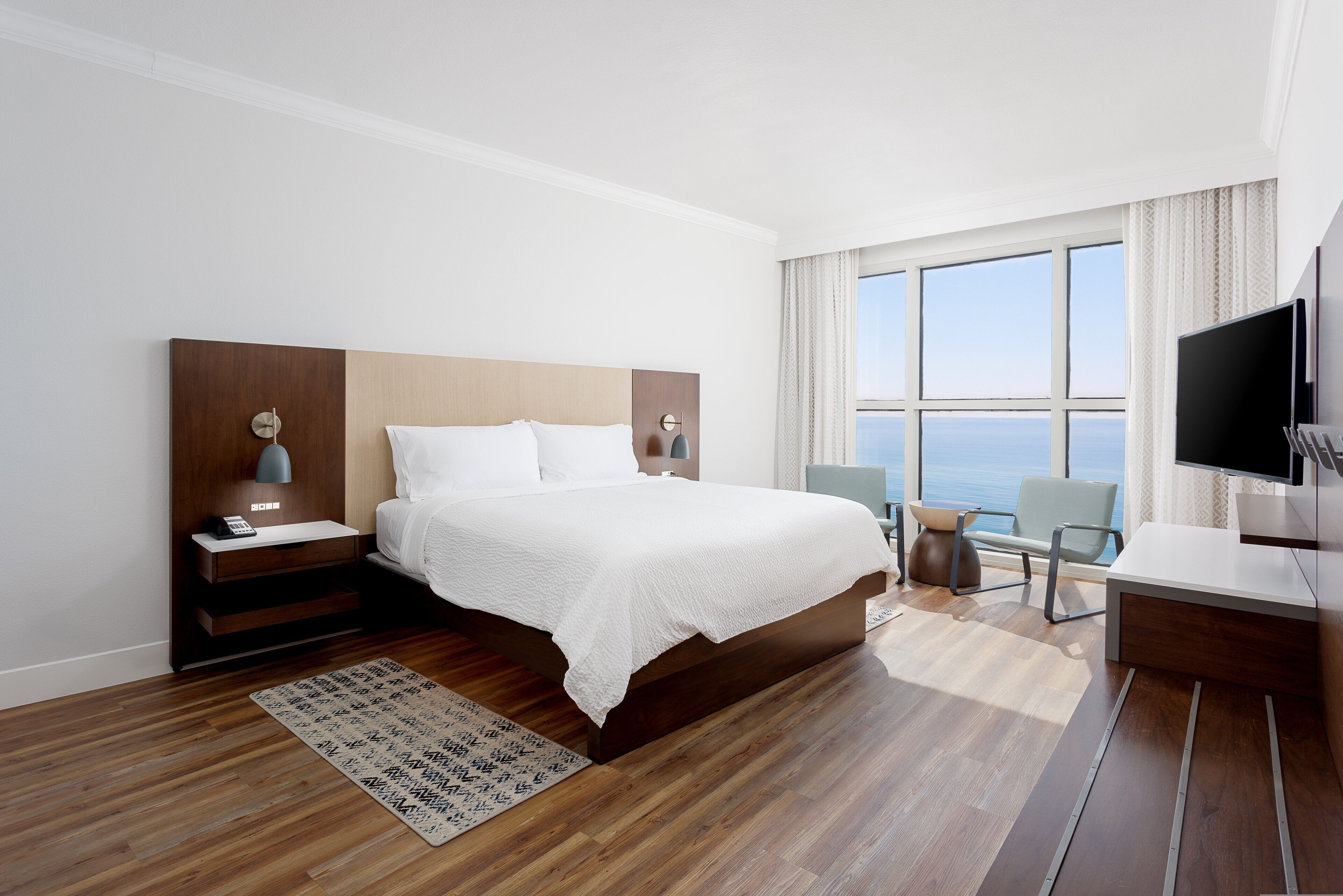 A modern hotel room with a large bed, bedside tables, a desk, a chair, and a TV.
