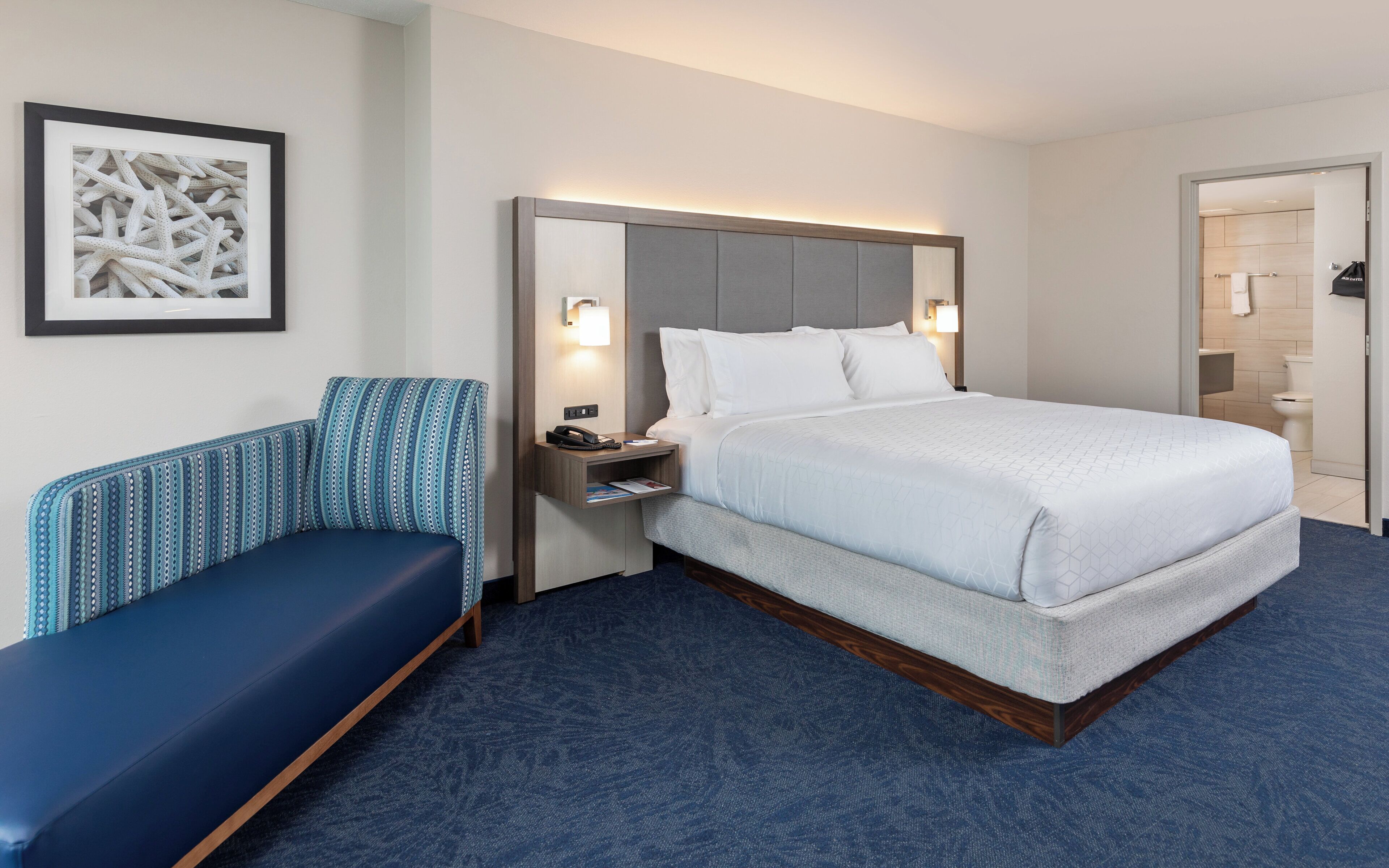 A hotel room with a bed, a bedside table with a lamp, a chair, and a framed picture on the wall.