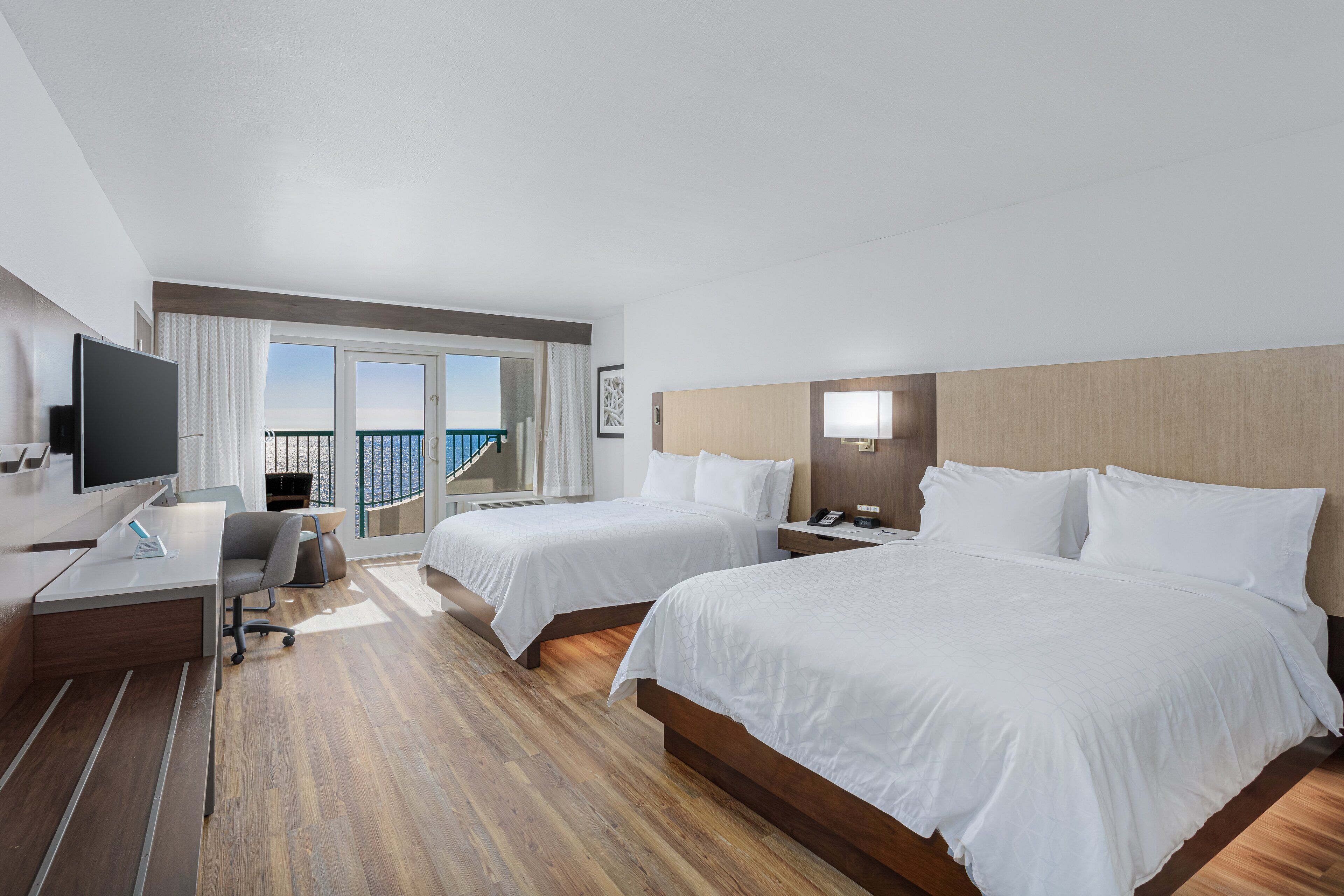 Ein Hotelzimmer mit zwei Betten, einem Schreibtisch mit Computer, einem Fernseher und einem Balkon mit Blick.