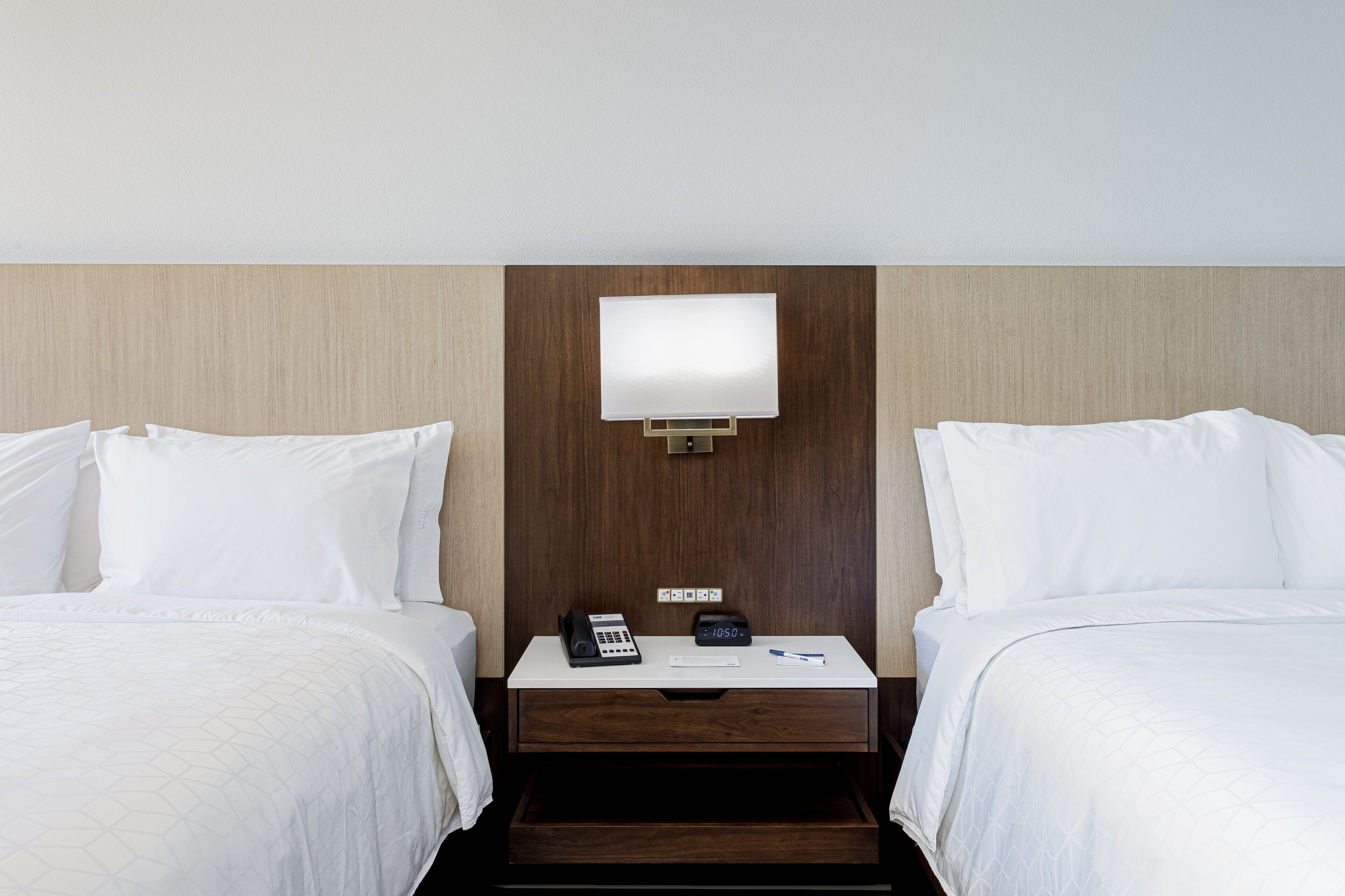Une chambre d’hôtel avec deux lits, une table de chevet avec un téléphone et une horloge, et une lampe fixée au mur.