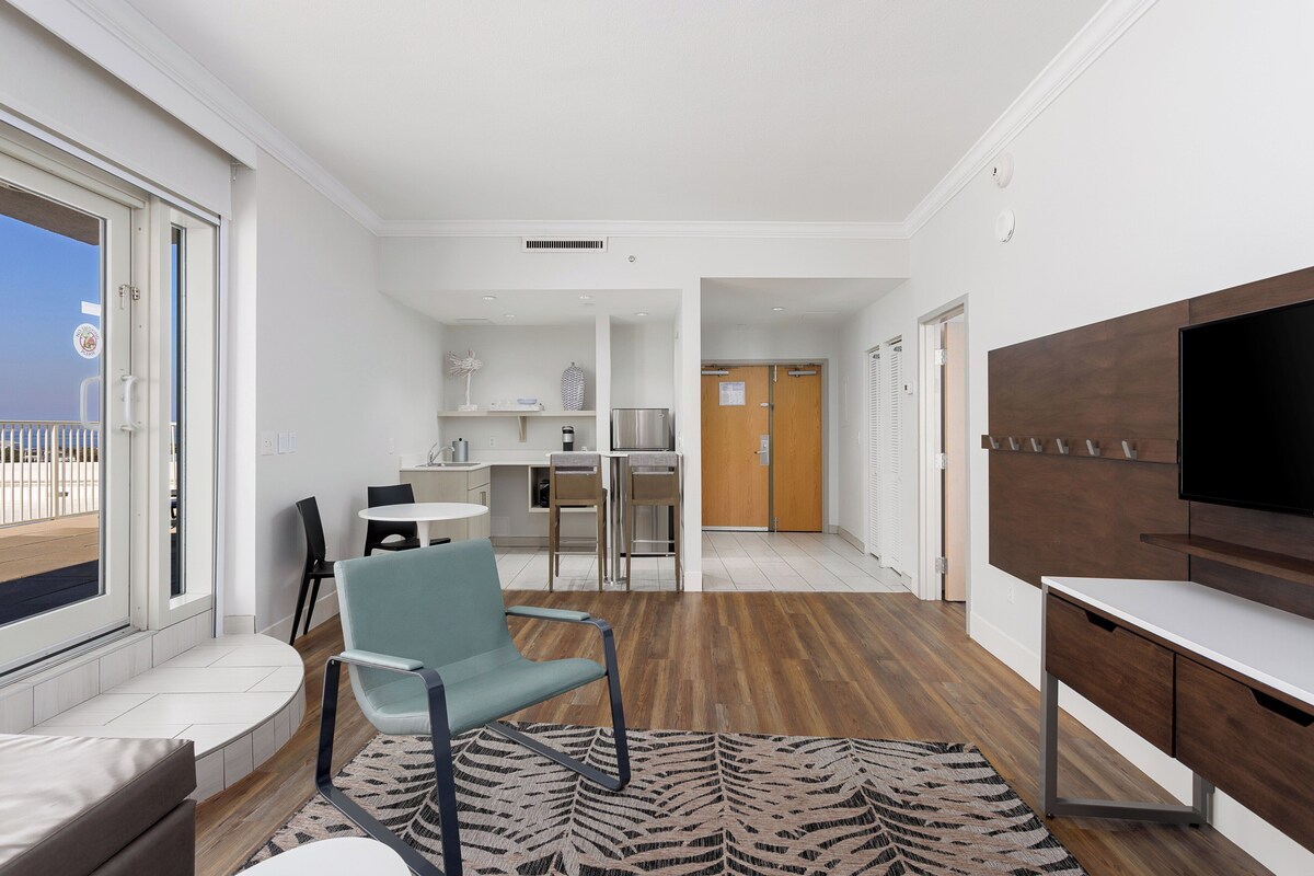 Habitación de hotel moderna con balcón, zona de comedor, televisor y suelo de madera.