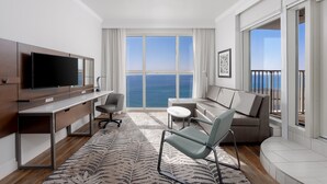 Una habitación de hotel moderna con un ventanal que ofrece vistas al mar, un televisor de pantalla plana, un escritorio, un sofá cómodo y una pequeña mesa redonda.