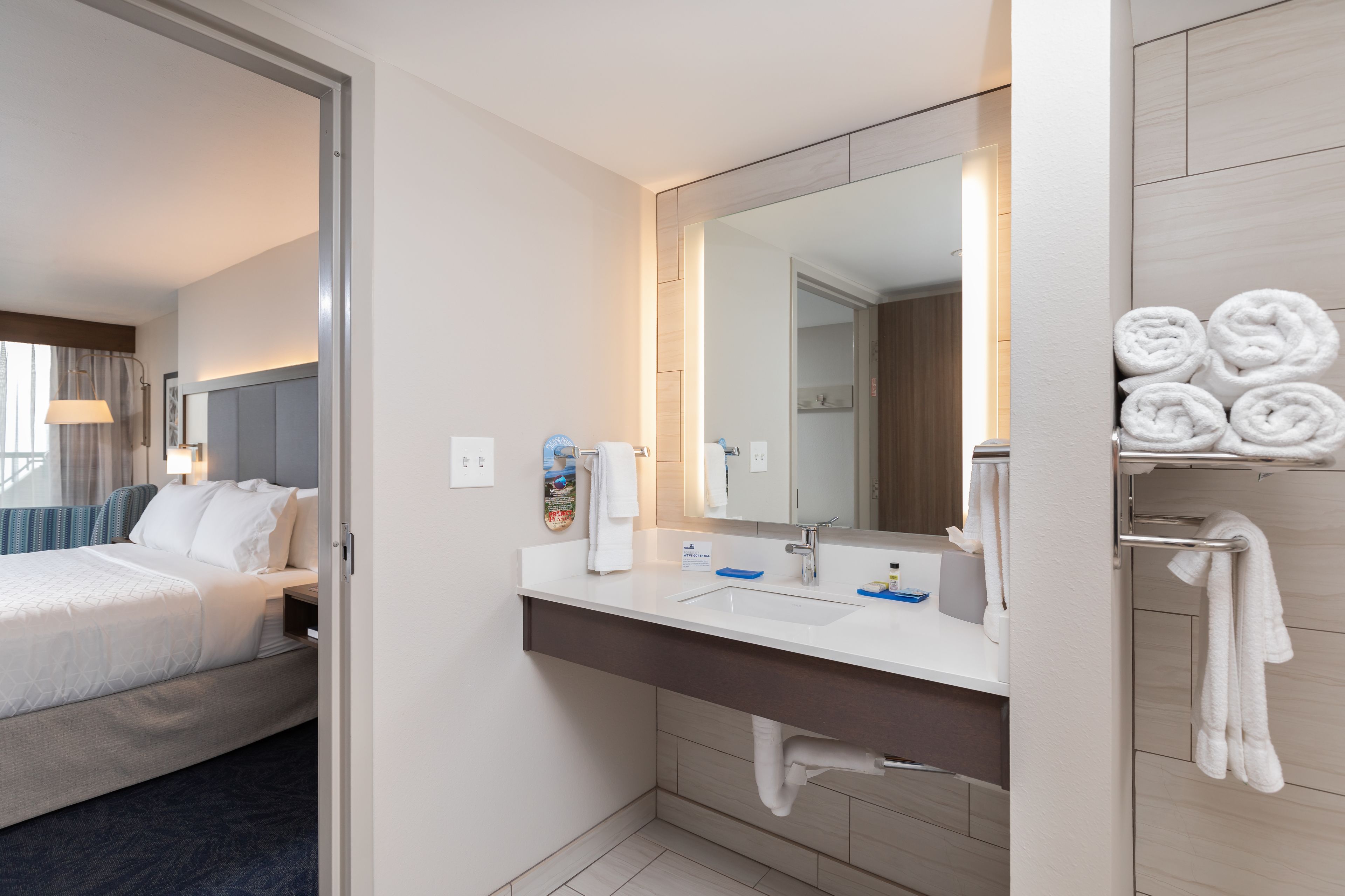 A hotel room with a bed, a bathroom with a sink, mirror, and towels.