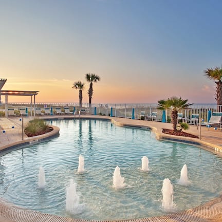 A pool with water fountains, palm trees, lounge chairs, and a view of the ocean at sunset.