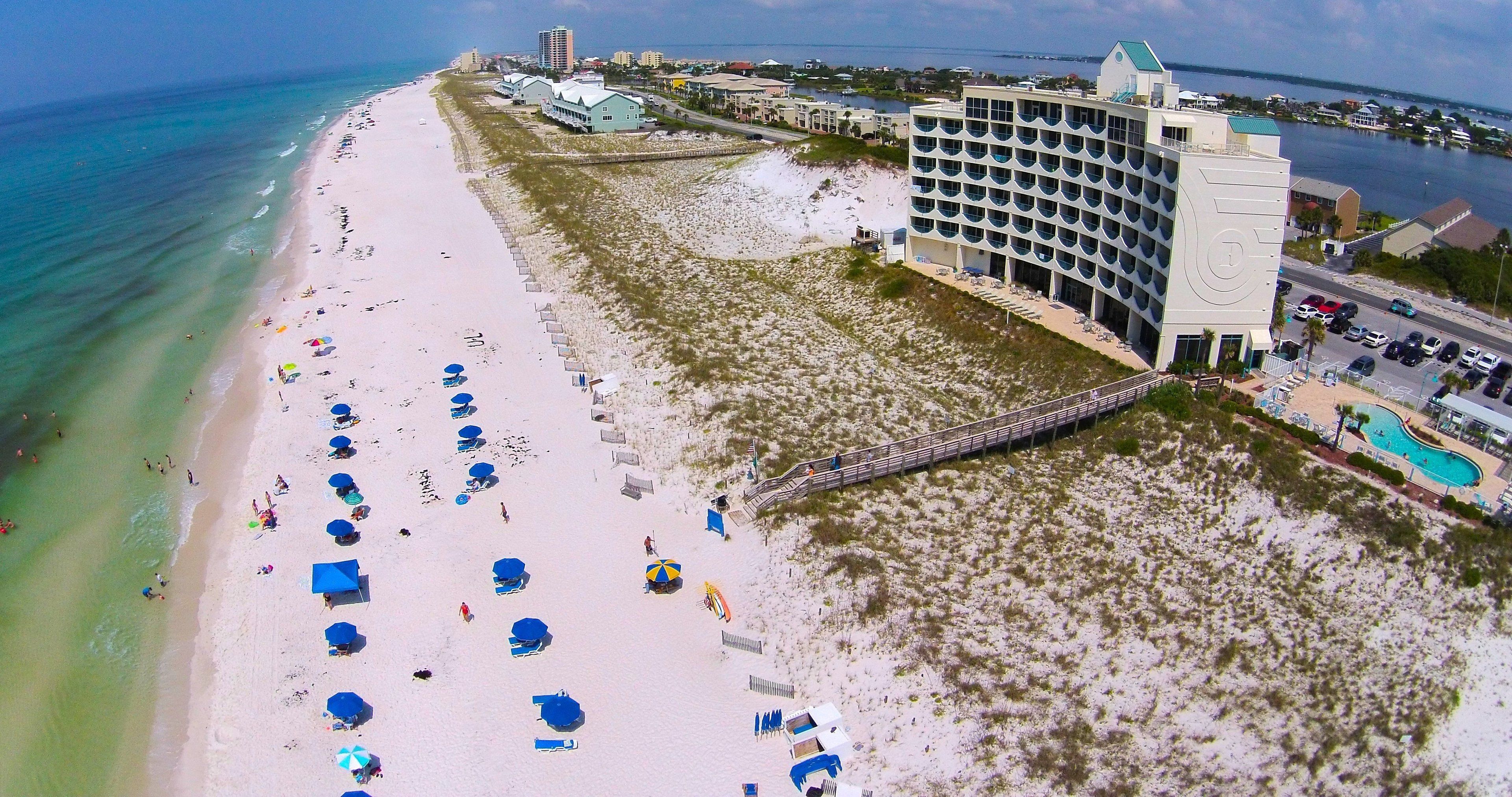 An aerial view of a beach resort with a large building, multiple beach umbrellas, and a swimming pool.