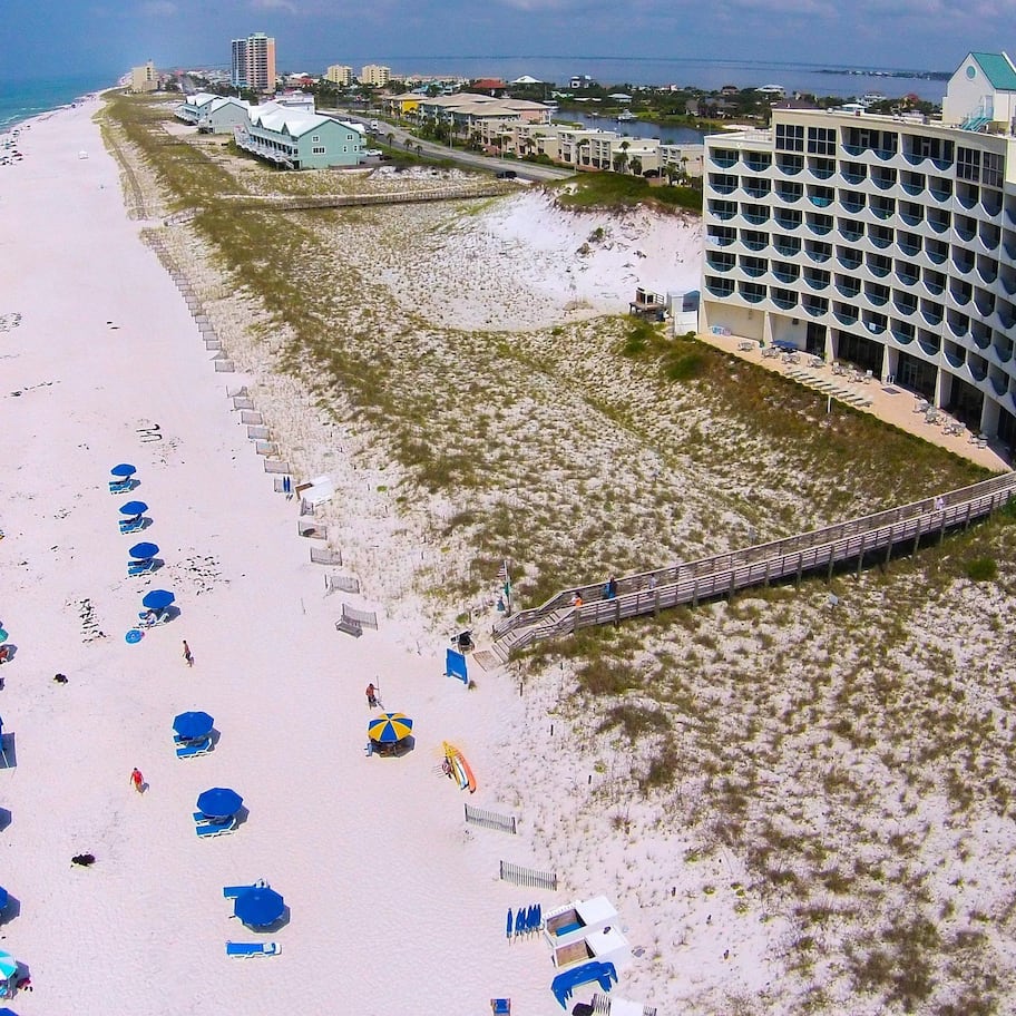 An aerial view of a beach resort with a large building, multiple beach umbrellas, and a swimming pool.
