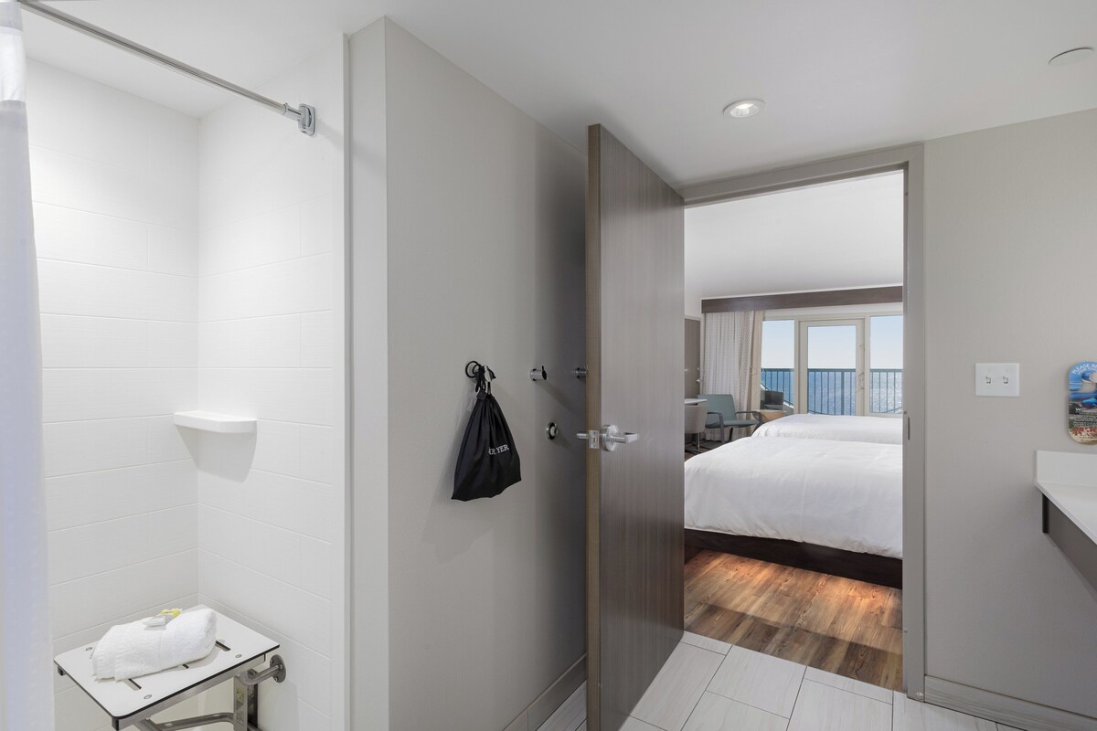 Habitación de hotel con ducha, una mesita con una toalla, una puerta que da a un balcón con cama y vistas.