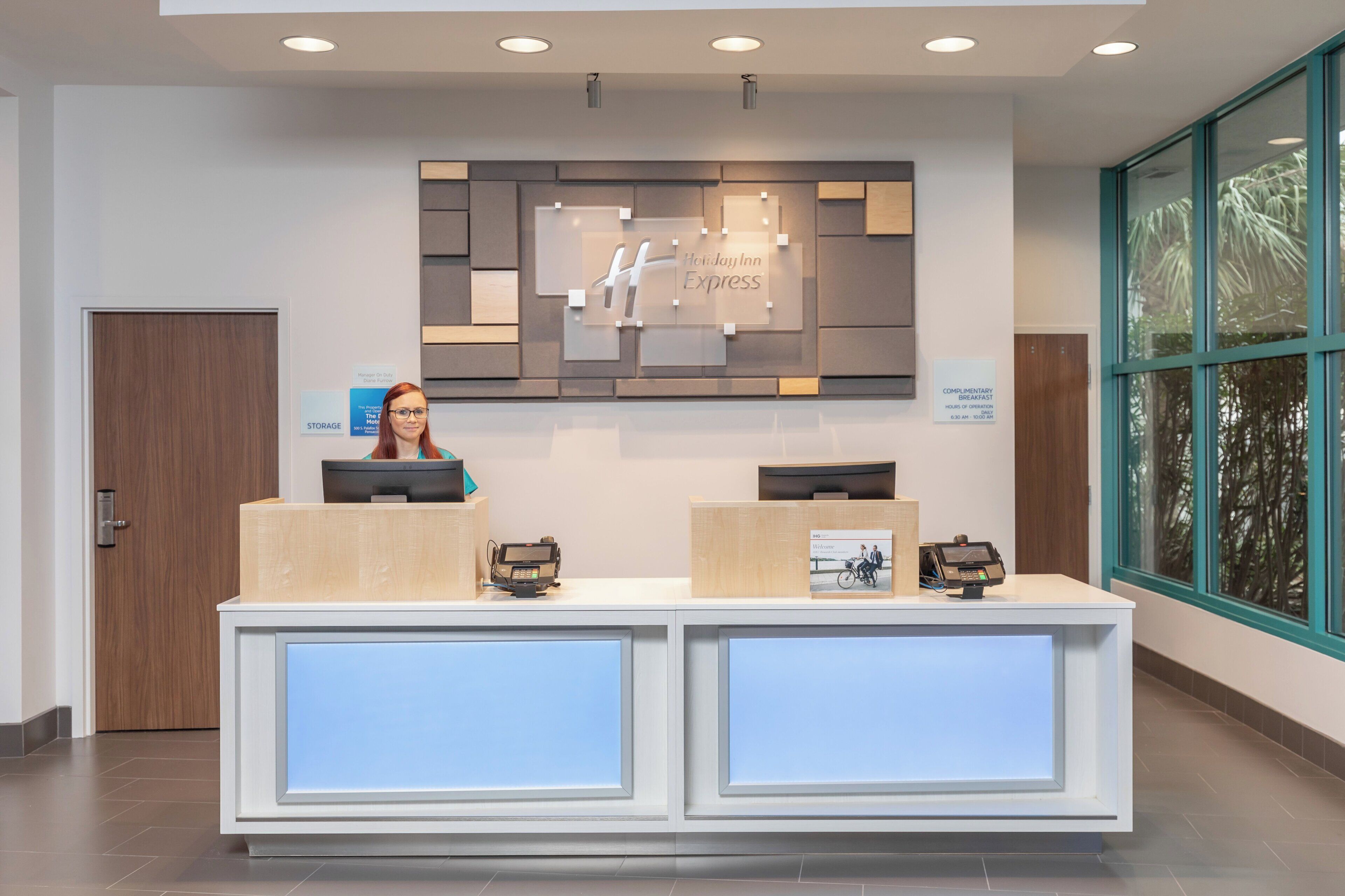A hotel reception desk with a digital display, a sign for complimentary breakfast, and a view of the outdoors through large windows.