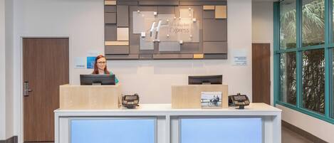 A hotel reception desk with a digital display, a sign for complimentary breakfast, and a view of the outdoors through large windows.