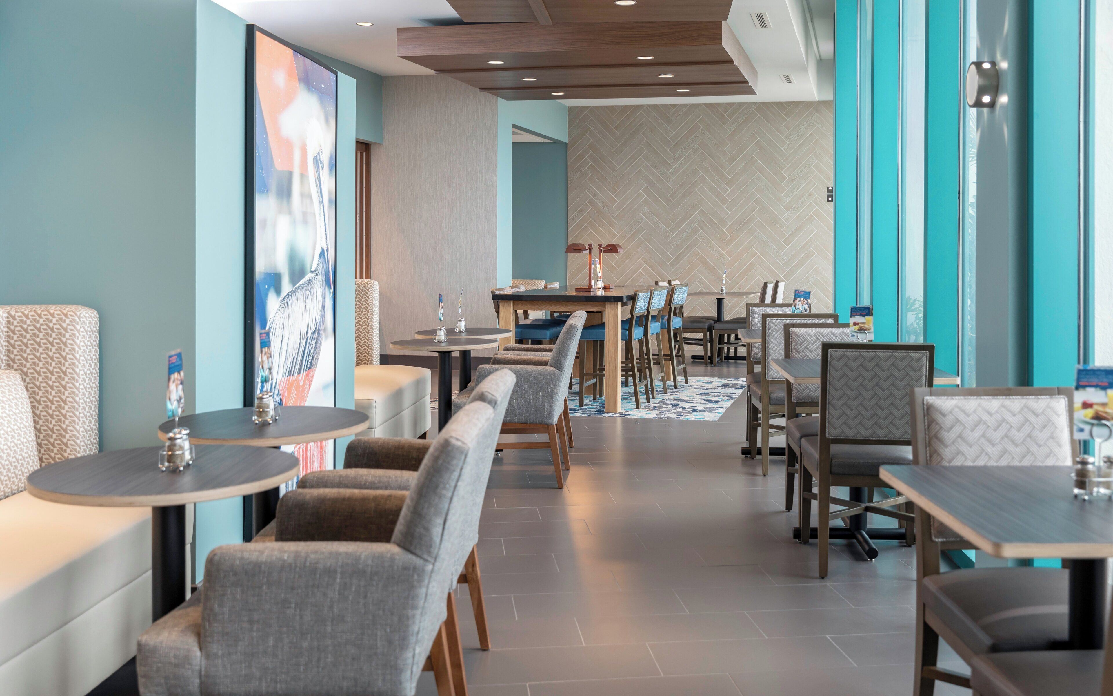 A modern restaurant interior with high tables and booths, a herringbone-patterned wall, and a large window.