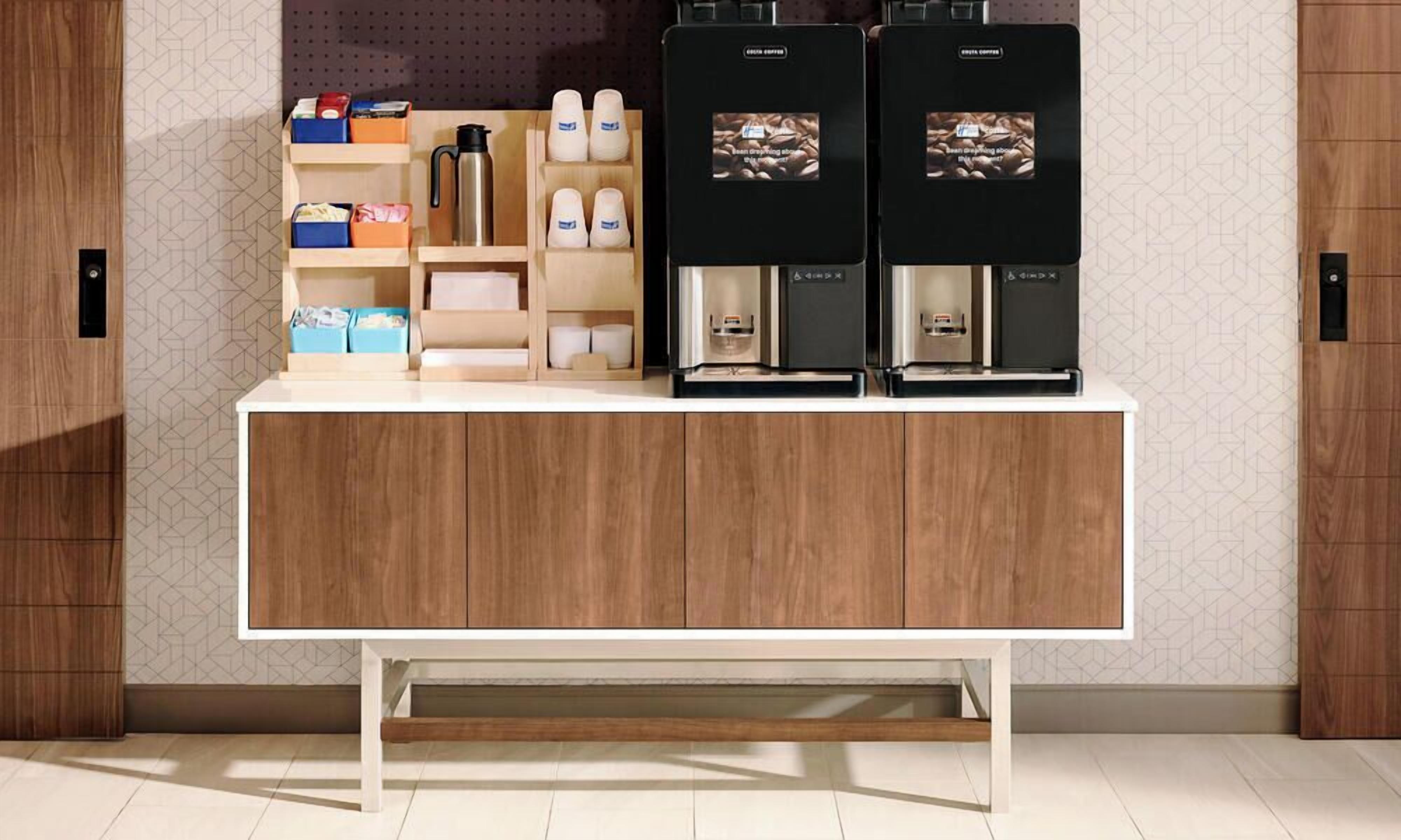 Una zona de cocina con encimera de madera, dos cafeteras y una estantería con diversos objetos, incluyendo cajas y botellas.