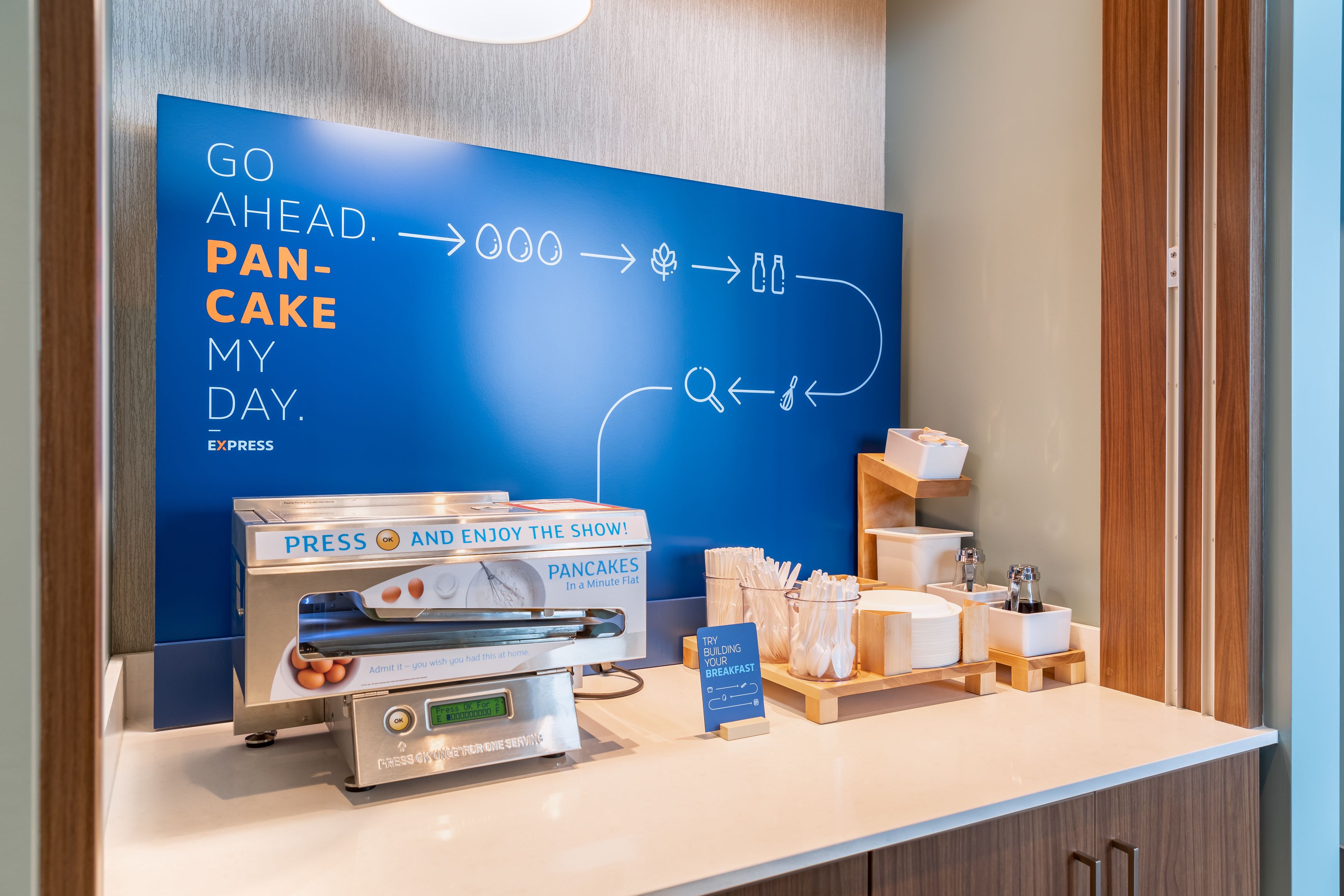 A breakfast station with a digital display, a pancake press, and a sign encouraging guests to try building their breakfast.